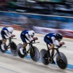 Três ciclistas competem lado a lado em uma pista coberta de velódromo.