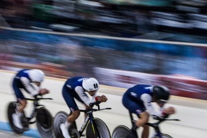 Três ciclistas competem lado a lado em uma pista coberta de velódromo.