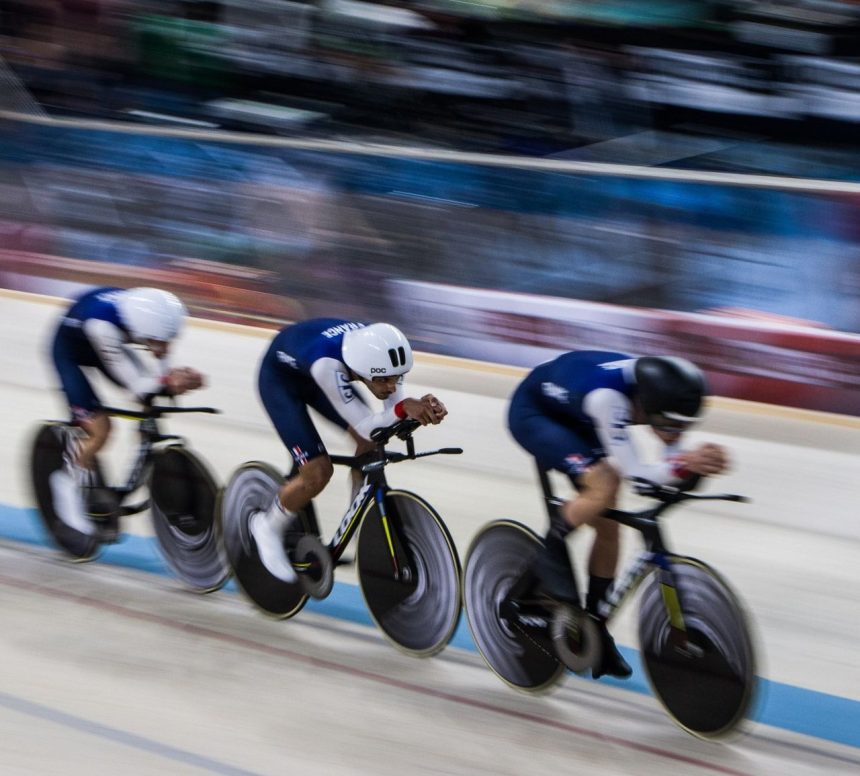 Três ciclistas competem lado a lado em uma pista coberta de velódromo.
