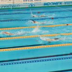 Nadadores disputam uma prova em piscina, avançando lado a lado pelas raias.