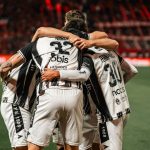 Jogadores de futebol com camisas listradas em preto e branco se abraçam e comemoram uma vitória no campo.