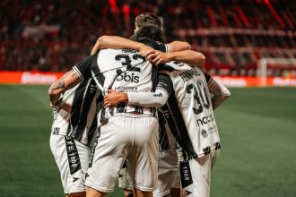 Jogadores de futebol com camisas listradas em preto e branco se abraçam e comemoram uma vitória no campo.