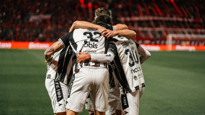 Jogadores de futebol com camisas listradas em preto e branco se abraçam e comemoram uma vitória no campo.