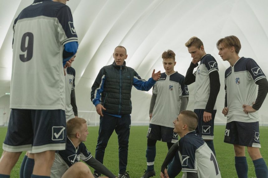 Treinador orienta com gestos uma equipe de futebol durante uma sessão de treino em um domo esportivo.