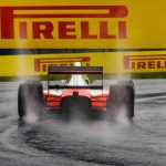 Carro de Fórmula 1 atravessa uma pista molhada, levantando spray sob uma placa da Pirelli.